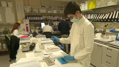 BBC A male scientist wearing a white lab coat and blue gloves is holding a pipette in one hand and a vial in another. In the background are other people sat looking at computer screens. There is a shelf filled with lots of folders on the wall.