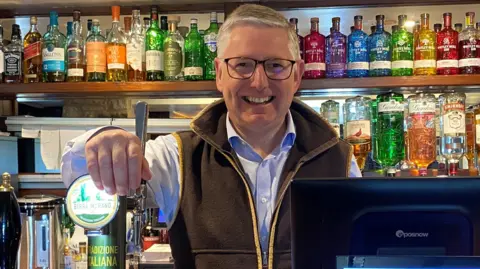 Scott Donaldson stands behind a pub bar with his hand on a beer pump. He's middle-aged, wearing a light blue shirt and brown sleeveless jacket and has glasses. Behind him are row upon row of spirits.