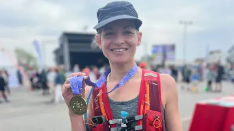 Family photo Carrie Jenkins after completing an ultra marathon - she is wearing a grey cap and a grey vest with a red running vest over the top for holding water and equipment. The back of her phone is poking out of the pocket on the right hand side of her chest,. Carries is holding a gold-coloured medal that says 'SheUltra, EST 2025' which is attached to a purple ribbon and she has it around her neck. The background is out of focus but you can see a crowd of people