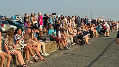 Dozens of people can be seen sitting on a small concrete sea wall at Crosby beach in Merseyside.