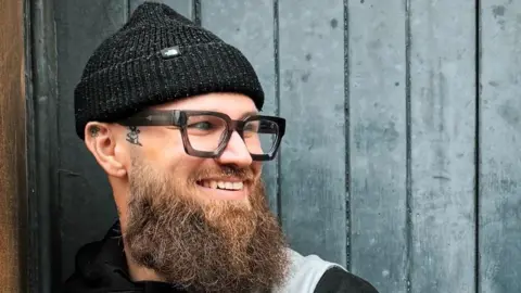 The Orchard Bristol A man wearing thick black glasses and a black beanie hat, with a big bushy brown beard, smiles in front of a black painted wooden door. 