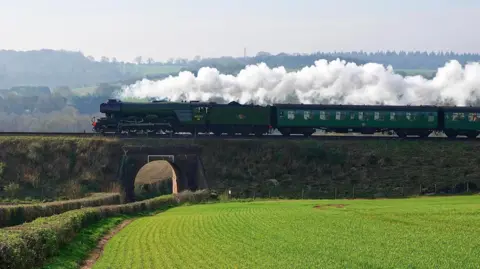 Soleil The steam train travels across the countryside and over a bridge.