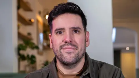 Bournemouth Foodbank Jon Saborido, a man of around 30-40 years old smilies warmly into the camera. He has short dark hair, brown eyes and is wearing a grey shirt with a collar and light brown t shirt underneath