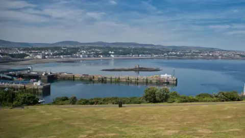 Manx Scenes A calm Douglas Bay featuring the Tower of Refuge on Conister Rock in the centre, with the green grass of Douglas Head in the foreground and Douglas Promenade in the distance in front of a a hilly landscape. The sky above is blue with thin while clouds.