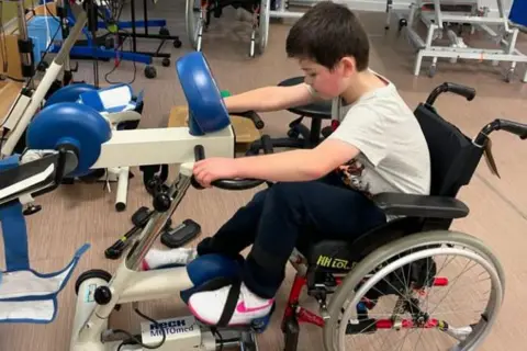 Family handout/PA Real Life Harry is seen in a wheelchair using a physical therapy exercise machine as part of a rehabilitation room with other equipment in the background. 