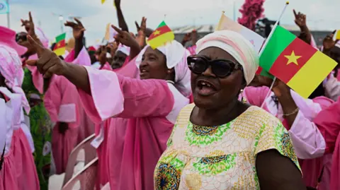 A woman wearing sunglasses, a headwrap and a patterned dress in cream, green and orange, smiling and cheering. Behind her, are women wearing pink, some of whom are pointing and others are waving Cameroonian flags