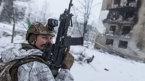A Ukrainian soldier wearing snow-camouflaged uniform holds a rifle pointing towards the sky and looks back towards the camera, as snow covers the land behind him and a building is seen badly damaged