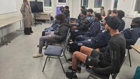 A group of young adults sitting on rows of chairs inside a classroom at community centre. There is a woman standing in front of the group.