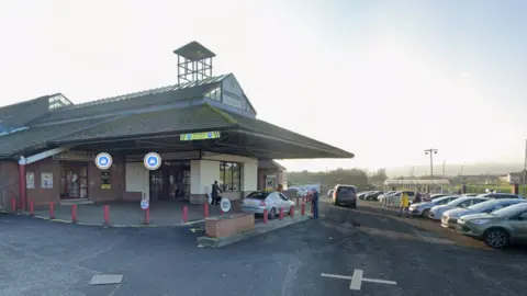 Exterior view of Northside Shopping Centre in Londonderry, showing the main entrance with a covered canopy, several people near the doors, and a car park with parked vehicles to the right on a bright, clear day.