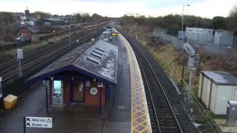 Two railway lines with a platform in between and a building and shelter on the platform. Buildings in the distance on either side of the line.