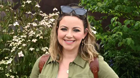 Charlotte Church is photographed smiling and looking at the camera, with flowers and greenery behind her. She has blonde shoulder length hair and sunglasses on top of her head. She is wearing a green long sleeve dress. 