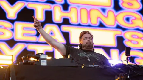 DJ Eats Everything performing on the stage. He is standing behind the decks and has one arm raised in the air. The screen behind him says 'EATS EVERYTHING' in yellow and pink writing.