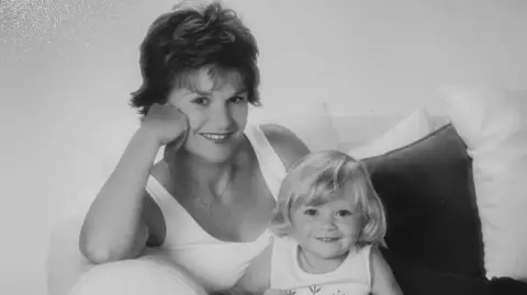 Daisy Daly Black-and-white portrait of Nicola and Daisy as a young child seated on a sofa. Nicola leans on an armrest with one hand supporting the head, wearing a sleeveless top and a short hairstyle. Daisy with light-colored, chin-length hair sits beside them, wearing a sleeveless outfit with a simple leaf design on the front. Both face the camera. Cushions and a plain wall form the background, with soft, even lighting across the scene.