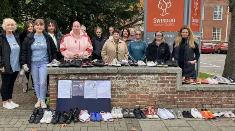A group of women are stood behind a wall which has rows of children's shoes lined up in pairs on it. Some of the shoes have a paper tag on them with a message. The are stood in front of a 'Swindon Borough Council' sign. 