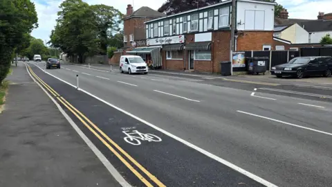 Paul Moseley/BBC A suburban street stretches into the distance, featuring a marked cycle lane separated by short bollards. Cars travel along the road while others are parked near a small parade of shops with flats on top.