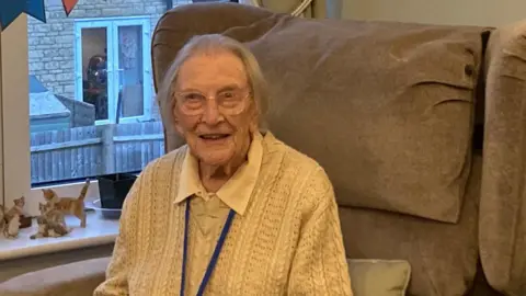 Jen Houston Mary sat in a comfy chair, smiling and wearing glasses. She has a cream wooly jumper on and a white blouse.