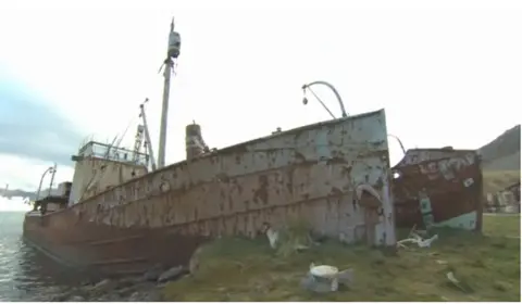 A rusty ship moored onto a remote shoreline with the hull at the forefront. It is grey and looks completely abandoned.