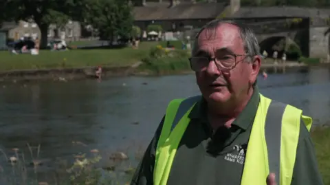 A man standing beside a river in a rural village setting on a sunny day is wearing a fluorescent yellow high-visibility vest with reflective strips and a dark green polo shirt.