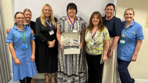Hospital staff posing for a picture. Two of the subjects are wearing blue uniforms. A woman in the centre of the image is holding a box containing equipment.