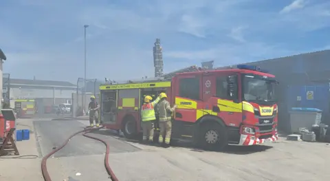 DWFRS Firefighters work next to a fire engine in a smoke-filled industrial yard