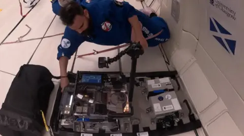 University of Glasgow Dr Gilles Bailet, wearing a blue flight suit, tests a 3D printer while strapped into a zero gravity aircraft. There is a saltire flag and the University of Glasgow logo taped to the wall of the aircraft.