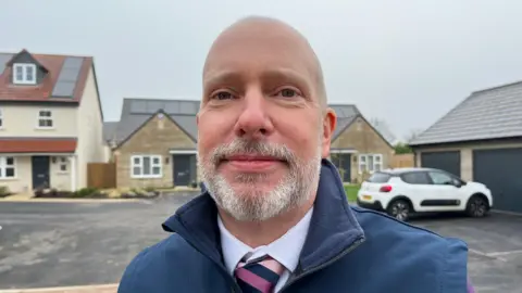 Mr Parker, wearing a white shirt, pink striped tie and blue zip up jacket. There are a number of new build homes with solar panels visible behind him.