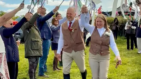 Safron and Nigel walking down a field with people either side holding decorations in the air. The pair wear black boots, beige trousers, brown tweed jacket and white shirts. Nigel holds Safron's hand into air.