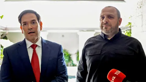 Reuters A man in blue suit, white shirt and red tie stands close to a man in a dark jacket. 