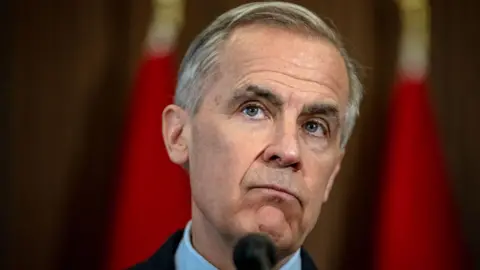 A close up image of Mark Carney, who looks out at the distance with a quizzical expression. Behind him is a blurred red background. He is standing at a podium. 