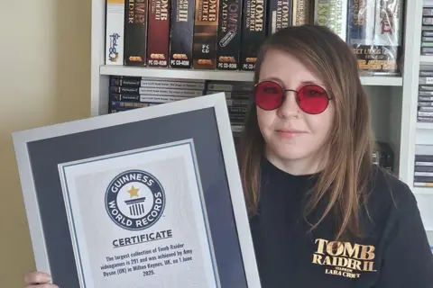 Amy Dyson A woman wearing red Lara Croft sunglasses and a T-shirt with "Tomb Raider 2" written on it is holding a frames Guinness World Record. She is stood in front of shelves stacked with video game boxes.