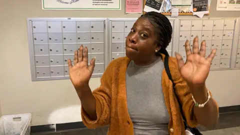 Woman in orange sweater with hands up in front of silver mailboxes