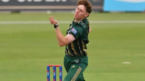 James Hayes, in green and gold kit, bowling alongside blue stumps with red bails