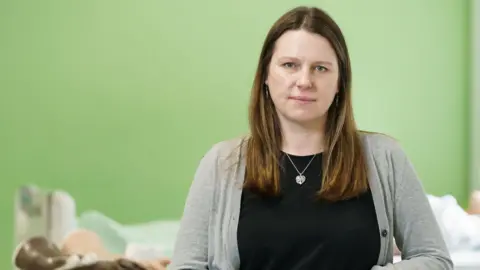 Dr Louise Harvey-Golding is in a room with a green wall in the background. She has long light-brown hair and blue eyes. She is wearing a grey cardigan over a black top and has a silver necklace with a heart-shaped pendant.