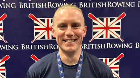 Ben Stephens A smiling Ben Stephens wearing his gold medal stands in front of the British Rowing logo.  