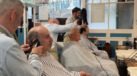 Three men sitting in chairs at the shop. They are getting their hair cut and dried and are covered by striped sheets. 