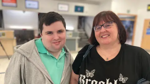 Thomas, who is has black hair and is wearing a turquoise polo shirt and a grey hoodie and is smiling at the camera, with Paula who is wearing a black T shirt and glasses. She is also smiling, they are standing in front of the baggage drop desk