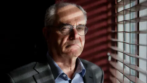 PA Media Clive Ruggles, who is looking serious and not smiling, is standing next to a window with wooden slatted blinds. He is wearing glasses and a grey suit with blue check shirt and has short white grey hair.