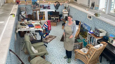 A woman with short grayish brown hair standing in a pool which is filled with various chairs, equipment and paintings. It's arranged so it creates an art studio.