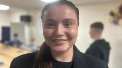 Zara is wearing a black suit jacket and black t-shirt. She has long brown hair tied back in a pony tail style. She is smiling at the camera. 