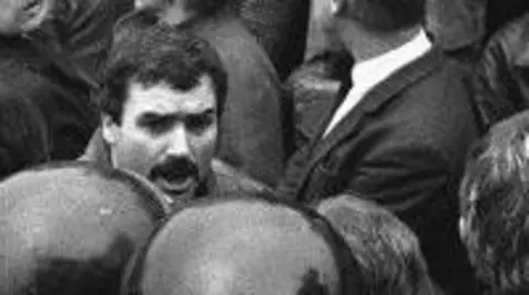 pacemaker Black and white photograph of a man in a crowd.
He has black hair and a black moustache
He is surrounded by men