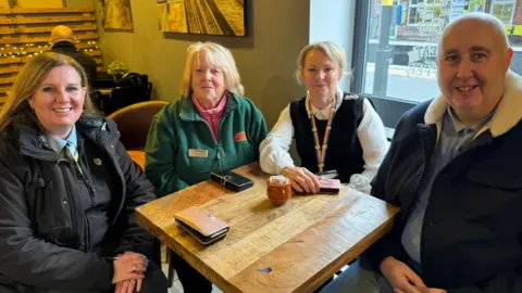 Sonya Harrison, Doreen Bird, Angela Horton en Ian Harrison zitten rond een houten tafel in café Coffee Carriage