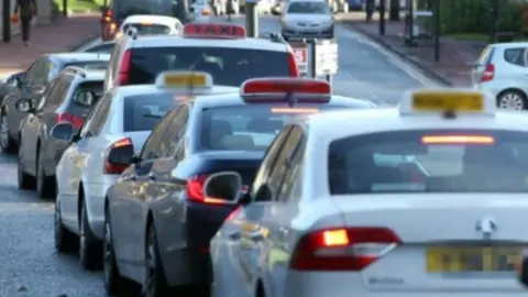 Taxis lined up in a street. Some are white, some black, and they have red or yellow signs on their roofs saying they are a taxi.