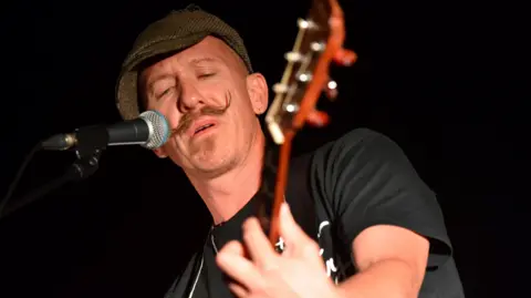 Foy Vance has a handlebar moustache and is wearing a tweed flat cap and a black short-sleeve t-shirt. He is holding a guitar out in front of him and his left hand is on the fretboard. To the right of his mouth is a microphone that he is singing into. 
