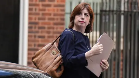 Lucy Powell is holding a brown folder and has a large brown-coloured bag over her right shoulder. She is wearing a dark blue jacket. A brick wall and a metal fence can be seen in the background.
