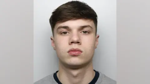 Wiltshire Police A mug shot of Aaron Nomicas. He has short dark hair and no facial hair. He is pictured against a white background, looking directly at the camera with a blank expression.