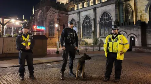 Three police officers standing in a row about a foot apart. The one in the middle is holding the lead of a police dog. They are stood on cobbled ground in front of a historic building