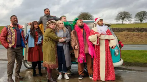 @SkyUK A group of people standing together in front of a turquoise and white camper van parked on a wet road. One person is dressed in a long red robe with white trim and is holding a white beard accessory. Others are wearing colourful winter clothing.