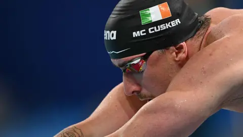 Max McCusker, wearing a cap with his country and name on it, prepares to enter the water during the 4x100m men's medley relay at the Paris Olympics