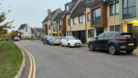 Elliot Deady/BBC A bus stop with three cars and one van parked across it. The bus stop is painted on the road outside a row of terraced houses.