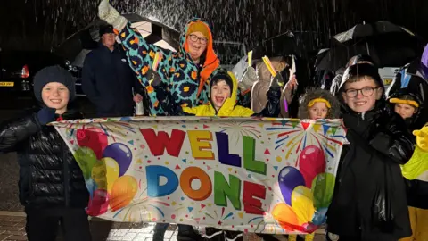 Four children and a woman stood outside with smiles on their faces. The children are holding a colourful banner with balloons on which reads 'Well done'.